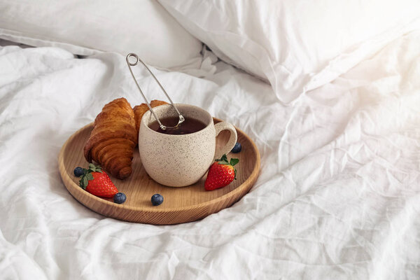 A cup of tea and croissants on the wooden tray standing on the bed with white bedsheets.