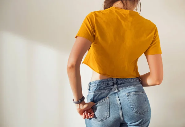 Jovem mulher vestindo camiseta amarela e jeans posando contra a parede branca, vista das costas — Fotografia de Stock