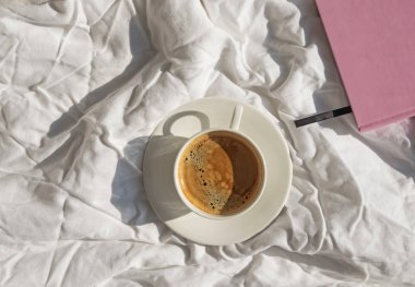 Sabah düz beyaz yatakta kahve, not defteri ve gözlük ile yatıyordu