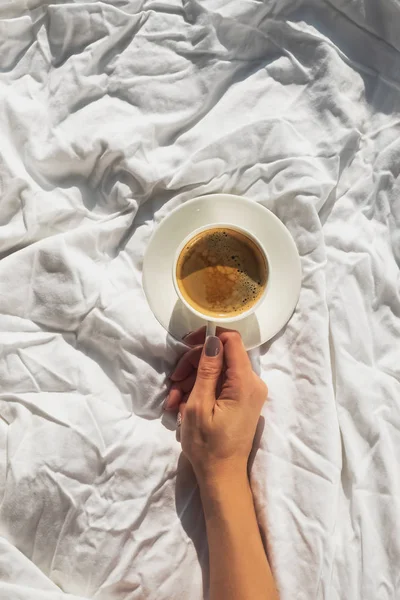 Kadın eli yatakta bir fincan kahve tutuyor. Odada beyaz çarşaf ve güneş ışığı var. — Stok fotoğraf