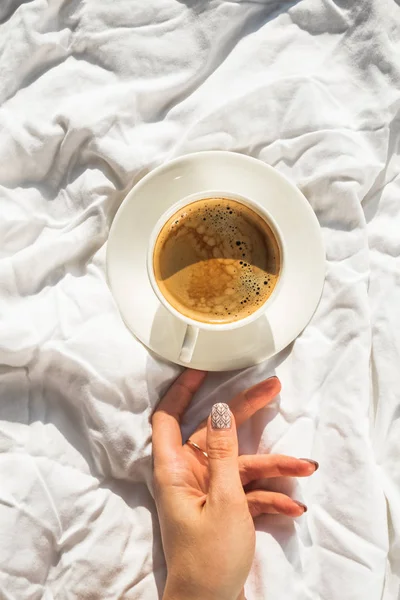Жінки рука тримає чашку кави на ліжку з білим полотном на кімнаті з природним сонячним світлом — стокове фото