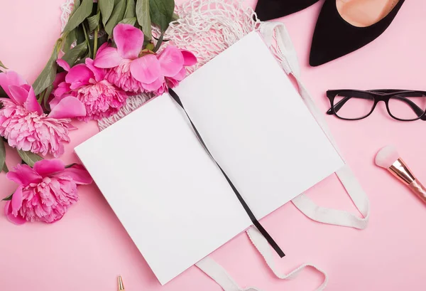 Mock-up de bloco de notas aberto na mesa com acessórios femininos e peônias rosa — Fotografia de Stock