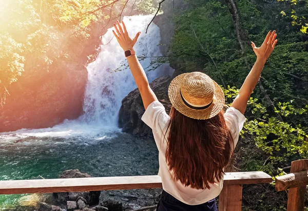 Jovem mulher de chapéu de palha em pé perto da cachoeira com as mãos para cima. E — Fotografia de Stock