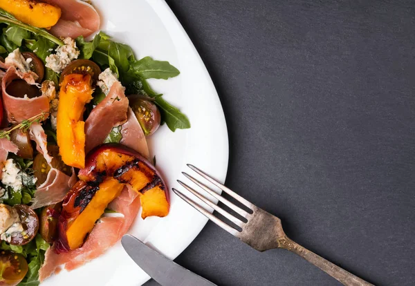 Köstlicher Salat mit gegrillten Pfirsichen und Prosciutto in einem weißen Teller auf schwarzem Schiefergrund — Stockfoto