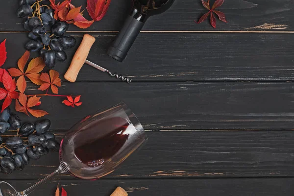 Winogrona, czerwone wino w butelce i jesienne liście na czarnym drewnianym stole — Zdjęcie stockowe