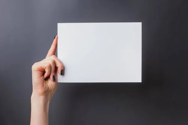 Womans hand with black nailpolish holding a blank paper against black wall