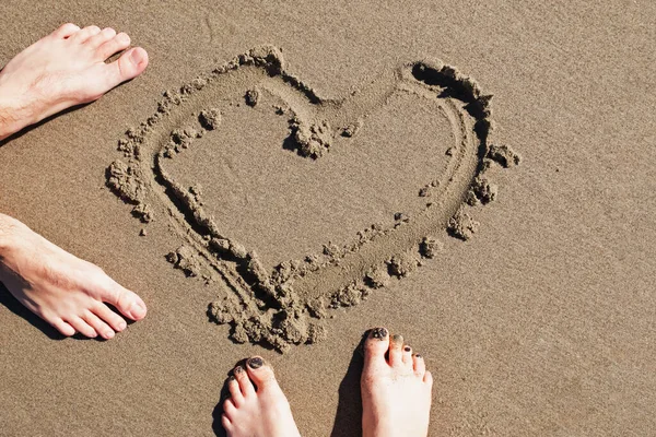 Füße von Mann und Frau, die auf dem Strand stehen und ein Herz in den Sand gezeichnet haben — Stockfoto