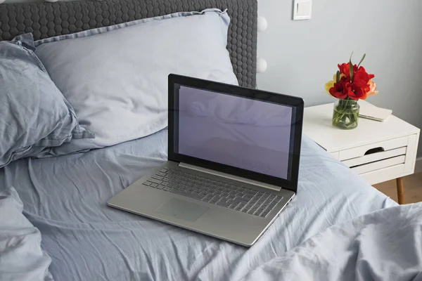 Puste, brudne, szare łóżko z laptopem w minimalistycznej sypialni z kwiatami na szafce nocnej — Zdjęcie stockowe