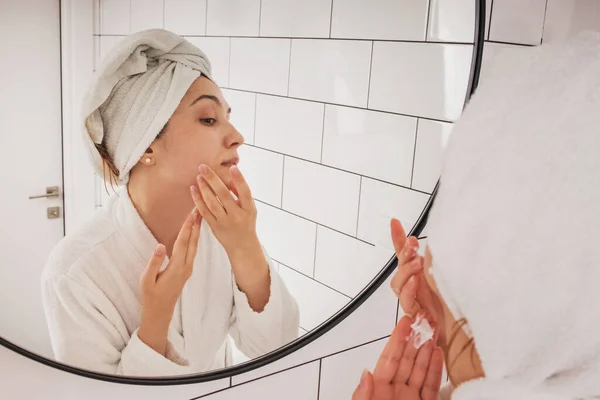Молода жінка наносить крем для зволоження на обличчя, стоячи у ванній — стокове фото