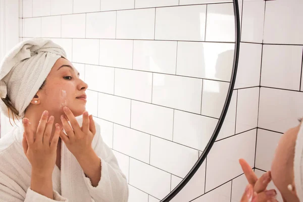 Young woman applying moisturizer cream on face standing in the bathroom — Stock Photo, Image