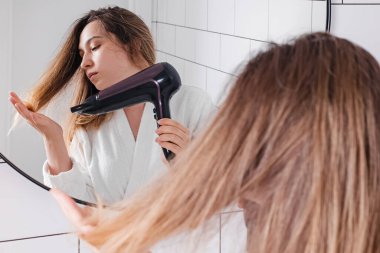 Genç ve güzel bir kadın banyoda saç kurutma makinesi kullanıyor.