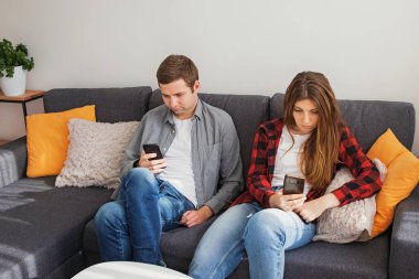 Genç çift, erkek ve kadın koltukta otururken cep telefonlarını kullanıyorlar.