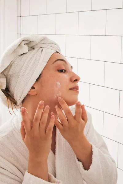 Young pretty girl applying moisturizer cream on face standing in the bathroom — Stock Photo, Image