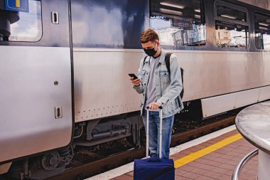 Trenin yanındaki tren istasyonunda duran koruyucu maskeli genç adam..