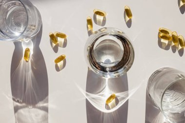Omega 3 kapsülleri ve beyaz masa üzerinde su olan bardaklar. Sert gölgeli yaratıcı kompozisyon. Sağlıklı yaşam tarzı.