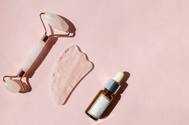 Yüz masajı için farklı araçlar ve pembe arka planda bir şişe nemlendirici serum. Dümdüz, sert gölgeli güneş ışığında yatıyordu..