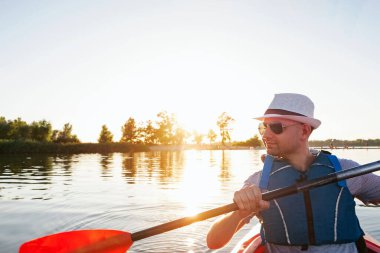 Şapkalı genç adam nehirde ya da gölde kano sürüyor.