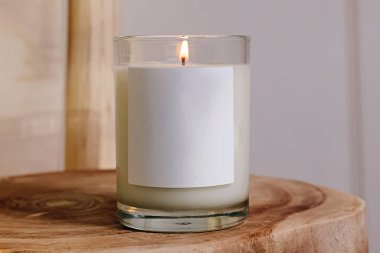 Cam bir kavanozda boş bir etiketle yanan beyaz bir mum, ahşap bir yüzeye oturmuş, sıcak ve sakin bir ortam yaratıyor..