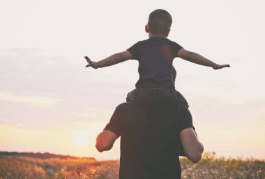 Oğlu babanın omuzlarında oturuyor ve onlar sahada gün batımına bakıyorlar.