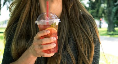 Kadın sokakta elinde pembe limonata olan plastik bardağı tutuyor.