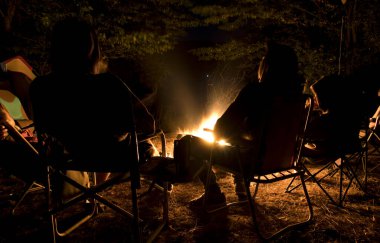 Gençler şenlik ateşinin etrafında oturup konuşuyorlar ve şarkılar söylüyorlar.