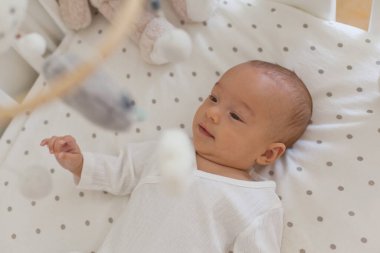 Cute 2-3 month old baby lying alone in a crib and playing with mobile toy above