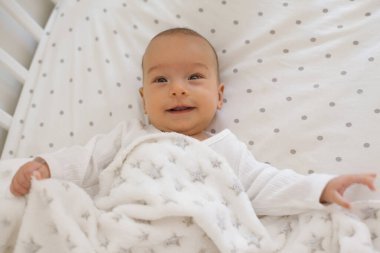 Smiling 2-3 month old baby lying on white bedding with soft fleece blanket, showing happy mood in the morning and looking at camera