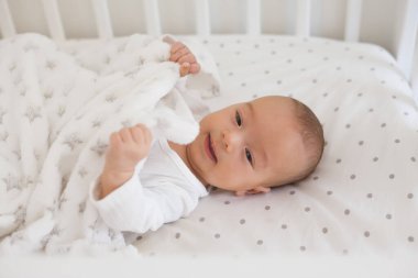 Smiling 2-3 month old baby lying on white bedding touching soft fleece blanket, showing happy mood in the morning