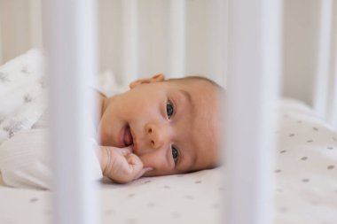 Smiling 2-3 month old baby lying on white bedding with soft fleece blanket sucking oma finger in mouth and looking at camera