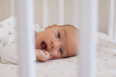 Smiling 2-3 month old baby lying on white bedding with soft fleece blanket sucking oma finger in open mouth and looking at camera