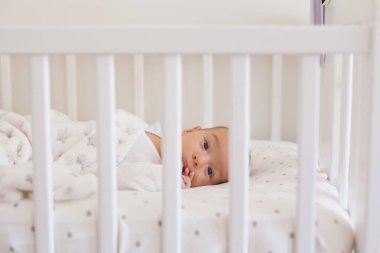 Smiling 2-3 month old baby lying on white bedding with soft fleece blanket sucking oma finger in mouth and looking at camera