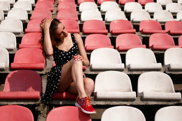 Young attractive woman seating the middle of an empty stadium. She is wearing a dress.
