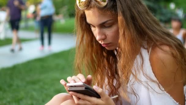 Retrato de uma jovem relaxada em um parque de verão lendo uma mensagem de texto em seu celular . — Vídeo de Stock