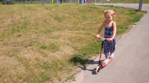 Маленька дівчинка швидко катається на скутері Кік вздовж шляху в літньому заміському парку. Повільний рух — стокове відео