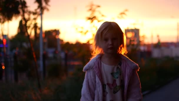 En liten blond flicka står i den nedgående solen strålar — Stockvideo