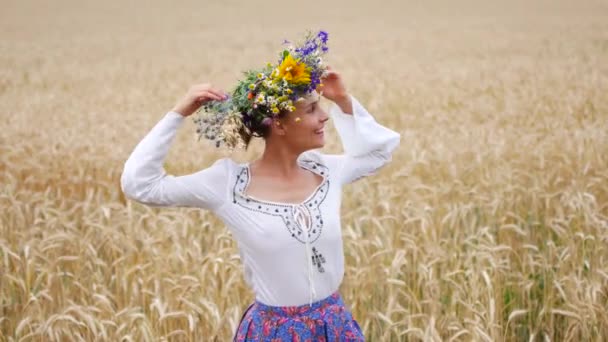 Felicidade, natureza, férias de verão, férias e conceito de pessoas - sorrindo jovem na grinalda de flores andando ao longo do campo de cereais — Vídeo de Stock