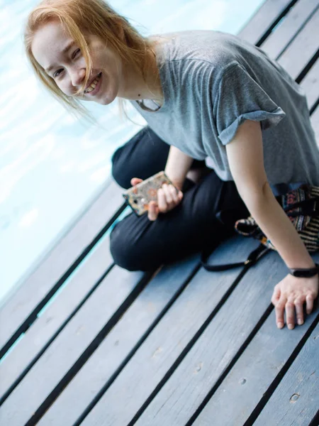 Portré, fiatal gyönyörű mosolygós szőke lány használ smartphone ülve a hídon. — Stock Fotó