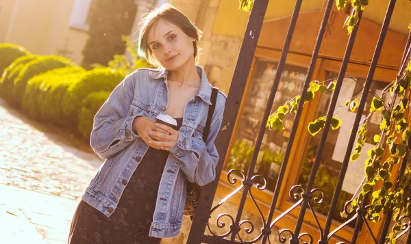 Uma mulher fica nos raios do sol poente com uma xícara de café. Retrato da cidade — Fotografia de Stock