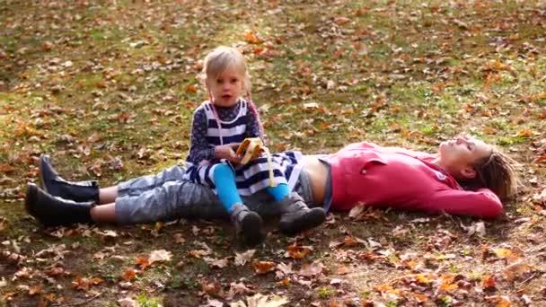 Hermosa mamá y lindo niño acostado en la hierba en el parque de otoño. Hija se sienta en mamá y come plátano, mamá mira al cielo — Vídeo de stock