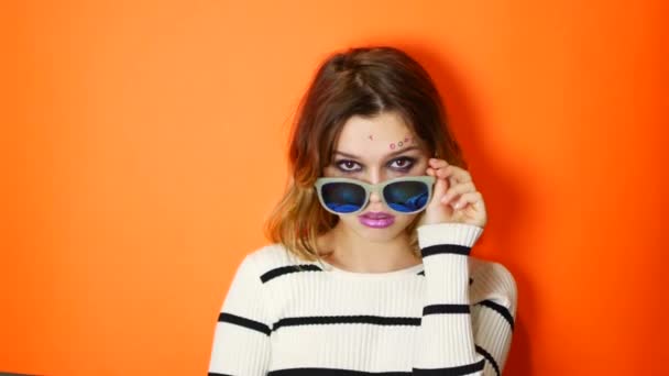 Скучная девушка в солнечных очках в руках на ярком оранжевом фоне в студии. Молодой высокомерный подросток — стоковое видео