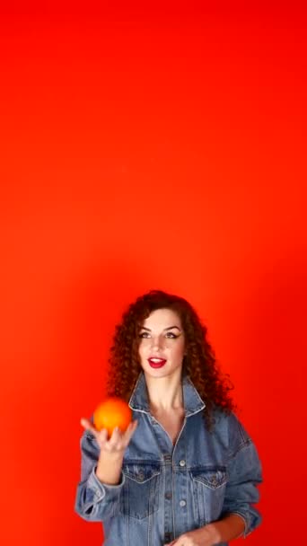 Молодая кудрявая девушка бросает апельсин на коралловый фон в студии — стоковое видео