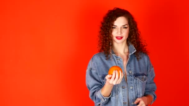 Menina encaracolado jovem joga laranja em um fundo de coral no estúdio — Vídeo de Stock
