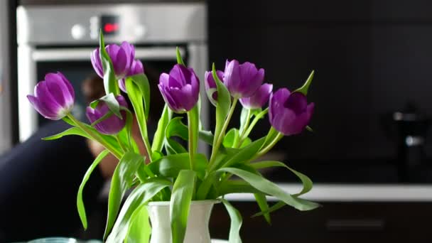 Un ramo de flores en un jarrón en la cocina. Contra el trasfondo en desenfoque, la mujer está ocupada con las tareas domésticas . — Vídeos de Stock