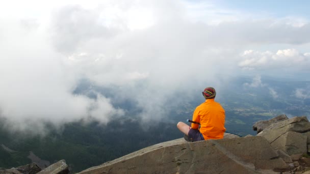 Caminante con una chaqueta brillante en la parte superior. Un turista mira las nubes y la vista desde la cima de la montaña . — Vídeos de Stock