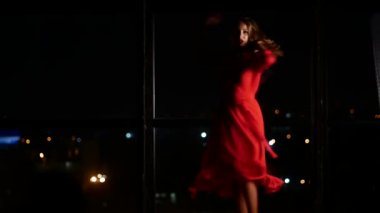 Kırmızı şık elbiseli bir kız akşam kenti olan büyük bir pencerenin önünde dans ediyor. Evde yalnız ve aşık.