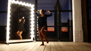 The girl slowly and sexually dances near a large mirror with bulbs