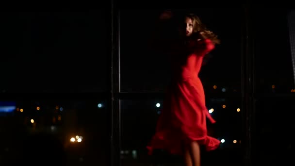Una ragazza in abito rosso elegante sta ballando sullo sfondo di una grande finestra con una città di sera. Da solo a casa e innamorato — Video Stock