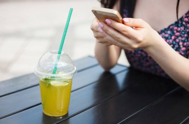 Genç kız, ellerinde smartphone aygıtı kullanırken ferahlatıcı soğuk mojito kokteyl içer. Her zaman bağlı kişi cep telefonu yeşil park gün kullanır. Soğuk içecek sıcak yaz günü tadını çıkarın.