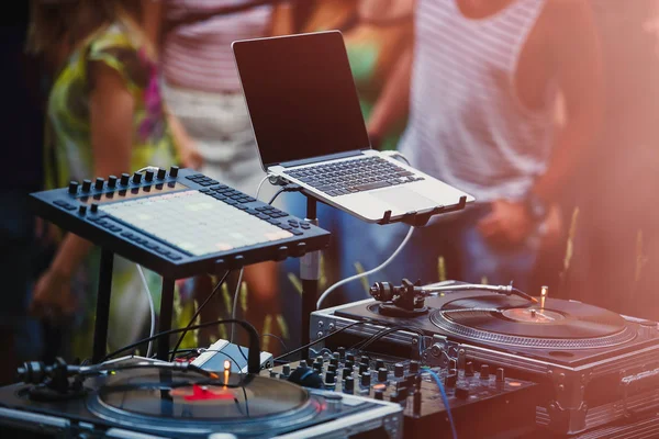 Open air music festival stage equipiment.DJ setup for playing music set on dance party.People dancing on background at backyard party.Disc jockey setup with midi controller & vinyl records player