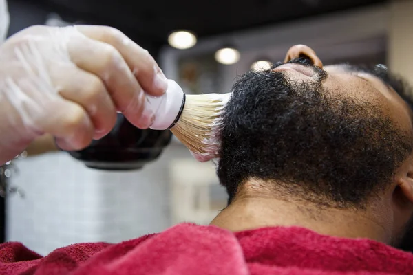 Barber hairdresser spreading white shaving cream with professional brush on black curly unshaven beard of african man client in barbershop. Male beauty treatment concept